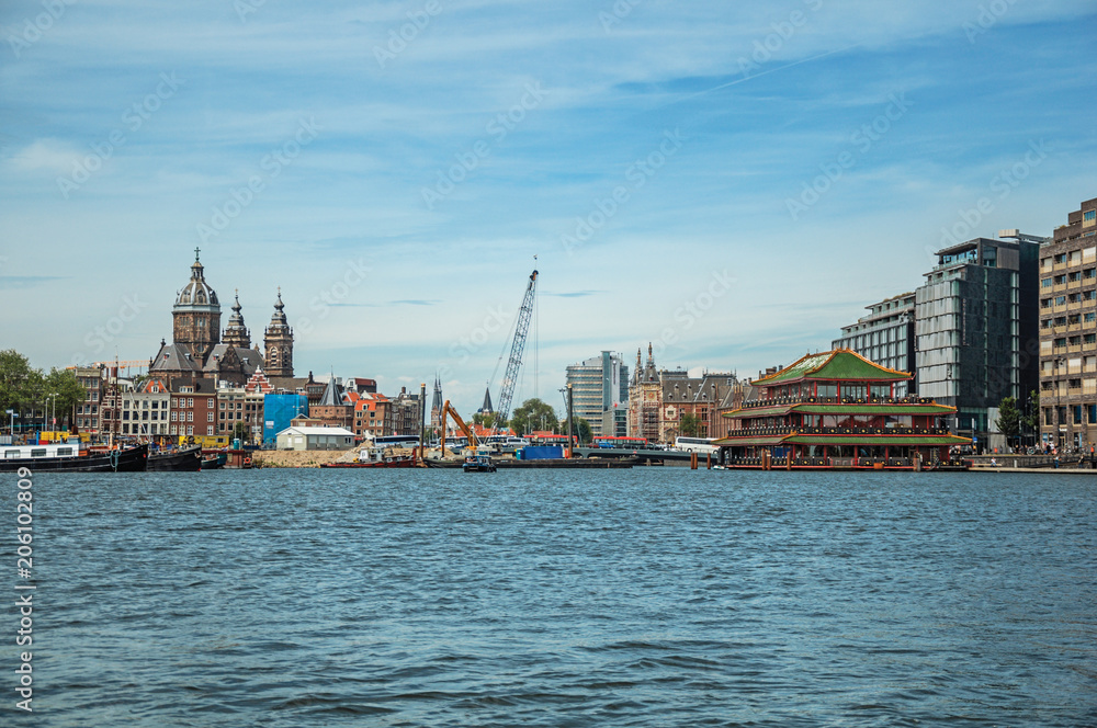 Fototapeta premium Port with ships, church towers and pagoda building restaurant on canal under a sunny blue sky in Amsterdam. Famous for its huge cultural activity, graceful canals and bridges. Northern Netherlands.