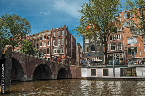 Bridge with bicycles, moored boats, old brick buildings and sunny blue sky in Amsterdam. The city is famous for its huge cultural activity, graceful canals and bridges. Northern Netherlands.