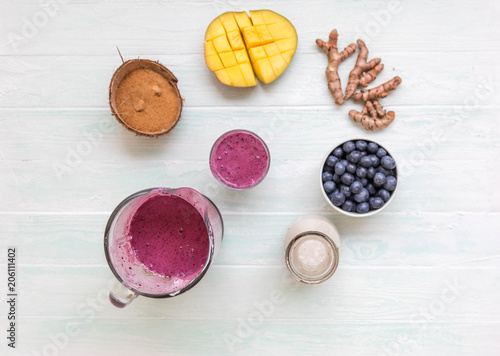 Mango coconut blueberry turmeric smoothie in dlass bowl with ingredients, coconut milk in glass bottle, coconut sugar in half coconut shells, berries,  turmeric root on light blue table,  photo