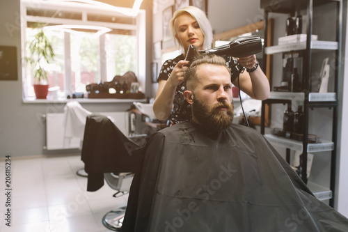 New hairstyle. Side view of young bearded man getting groomed at hairdresser with hair dryer while sitting in chair at barbershop.