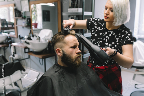 New hairstyle. Side view of young bearded man getting groomed at hairdresser with hair dryer while sitting in chair at barbershop.