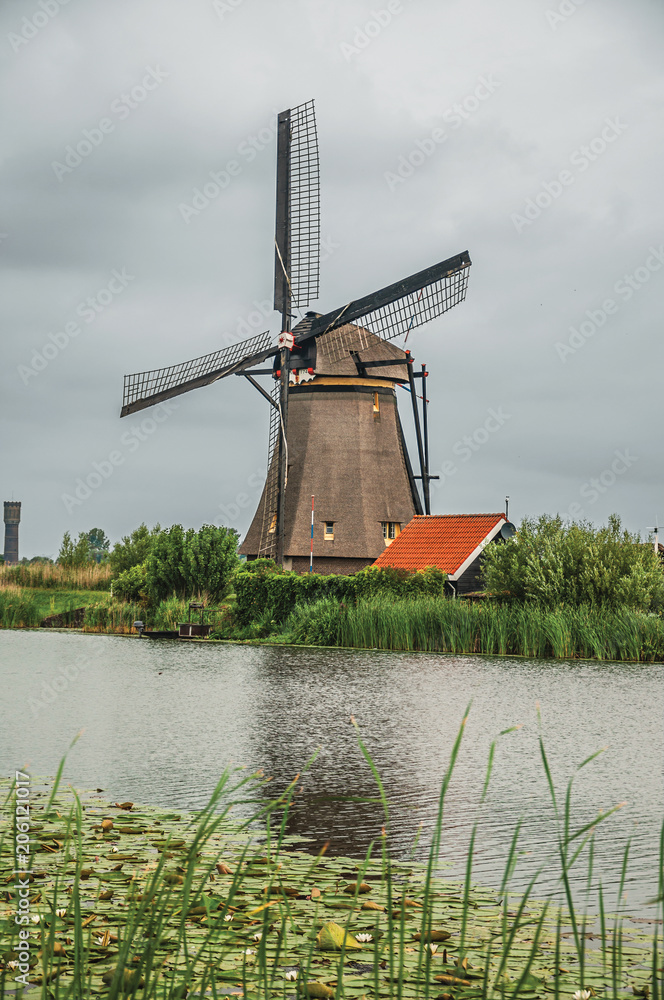 Windmill with house and bushes on the bank of a large canal in a cloudy day at Kinderdijk. Situated in a polder, has the largest concentration of old windmills in the country. Southern Netherlands.