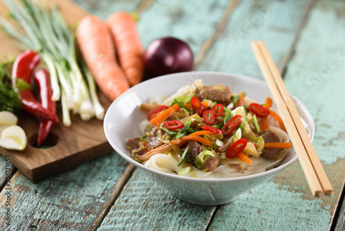 Tasty traditional Asian noodles with meat and vegetables in white bowl on rustic blue wooden background