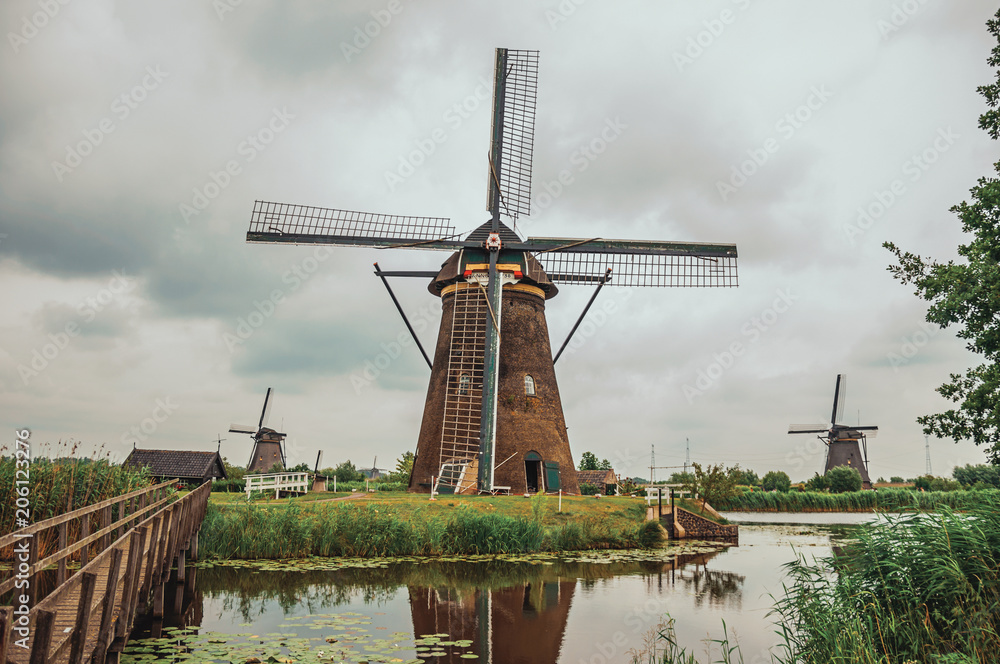 Windmills and bushes on the bank of a large canal in a cloudy day at Kinderdijk. Situated in a polder, has the largest concentration of old windmills in the country. Southern Netherlands.