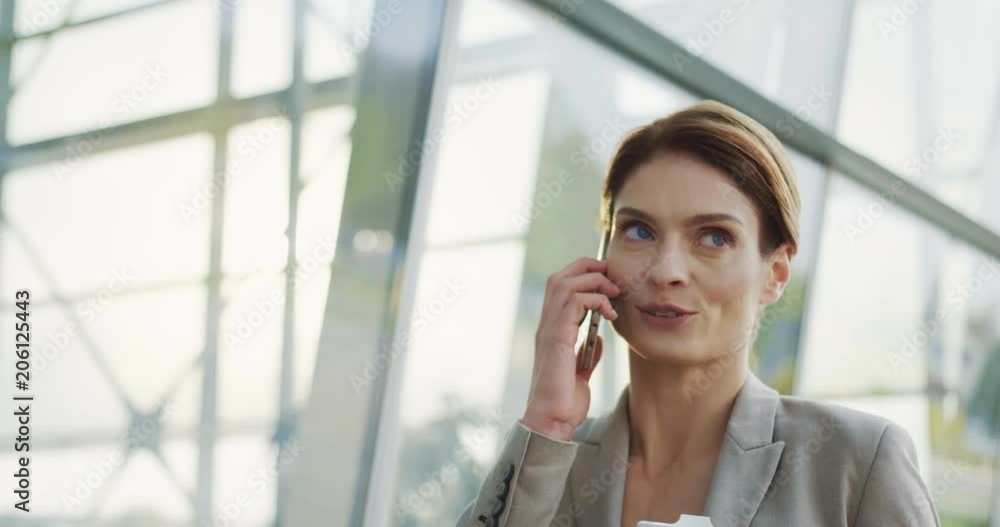 Close up of the beautiful smiled woman walking the office center passage, talking on the phone and carring her morning coffee. Sunny day. Close up of the beautiful smiled woman walking the office center passage, talking on the phone and carring her morning coffee. Sunny day.