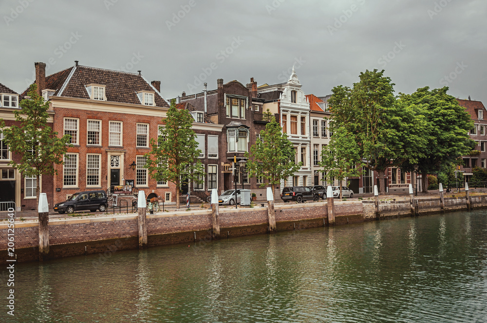 Fototapeta premium Wide tree-lined canal with brick buildings in a street next to it and cars in a cloudy day at Dordrecht. Important and historic port city bordered by rivers. Southern Netherlands.