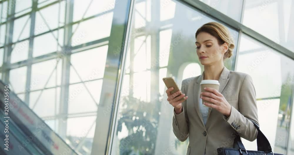 Caucasian pretty businesswoman in the suit walking in the office center glass passage, taping on the phone and carrying her morning coffee on the sunny day.