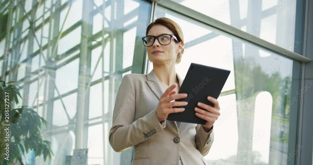 Portrait shot of the Caucasian pretty business lady taping on the tablet computer, smiling and looking at the side while posing on the glass building background. Outdoor Portrait shot of the Caucasian pretty business lady taping on the tablet computer, smiling and looking at the side while posing on the glass building background. Outdoor