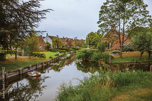 Foto Charming small canal next to rustic houses with lush gardens and trees reflected on the water in cloudy day at Drimmelen