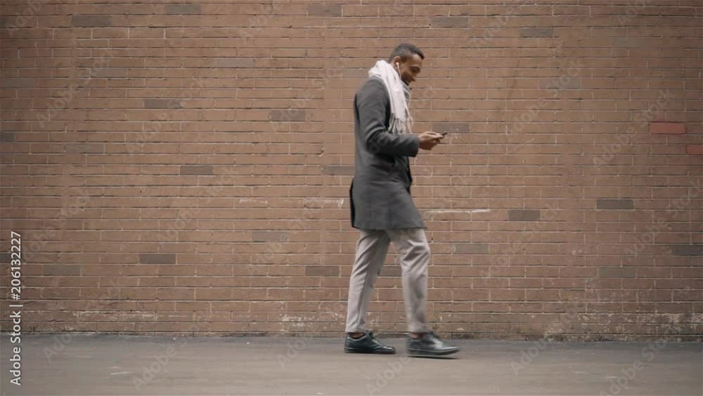 African American man in a coat and scarf listening to music and walking along a brick wall on an autumn day. Tracking slow motion medium shot African American man in a coat and scarf listening to music and walking along a brick wall on an autumn day. Tracking slow motion medium shot