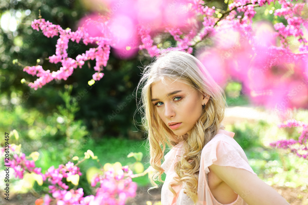 Fototapeta premium Beautiful girl standing under gentle tree with tiny hot pink blossom. Charming blond young woman breathing fresh aroma of blooming flowers outdoors, spring time concept