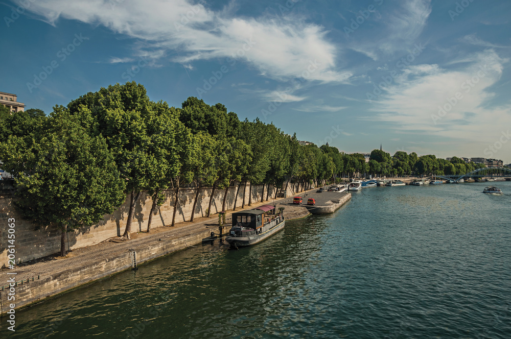 Seine River bank with walls, greenery and boats under sunny blue sky in Paris. Known as the “City of Light”, is one of the most impressive world’s cultural center. Northern France. Seine River bank with walls, greenery and boats under sunny blue sky in Paris. Known as the “City of Light”, is one of the most impressive world’s cultural center. Northern France.