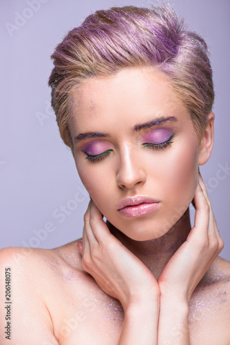 attractive woman with violet glitter on neck and short hair touching neck with closed eyes isolated on violet