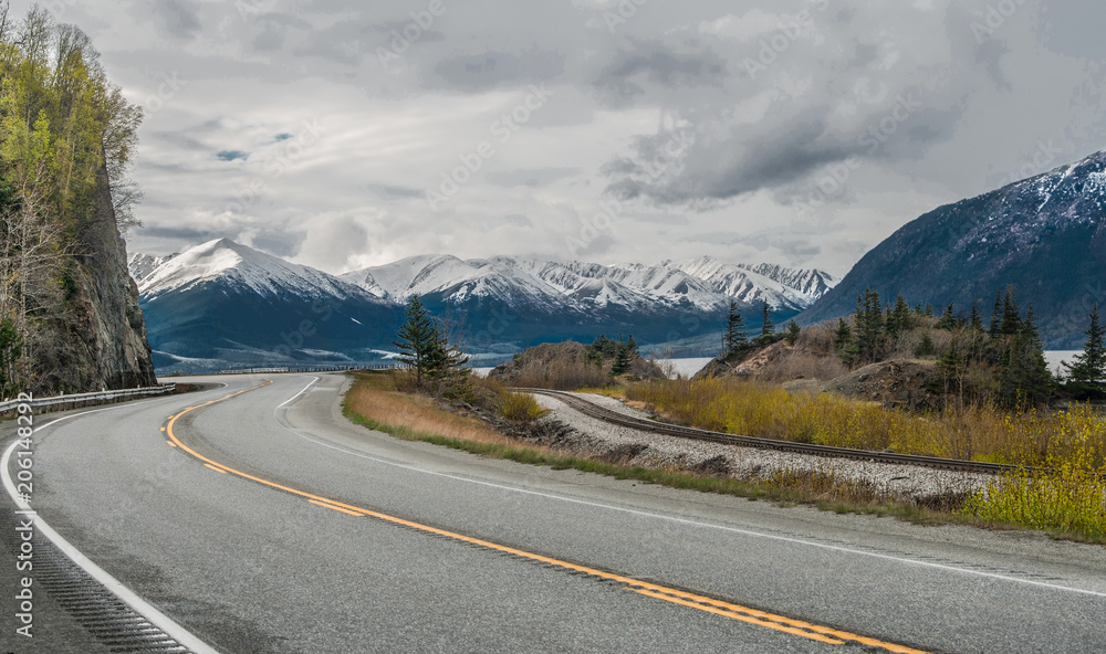 Alaska Scenic Road:  The Seward Highway curves beneath cloudy skies as it passes by snow-covered mountains at the edge of an ocean inlet south of Anchorage.