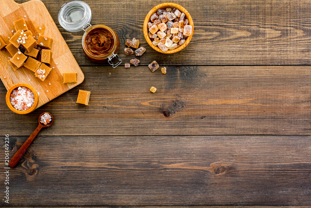Cook caramel sauce. Melted caramel in glass jar, caramel cubes on cutting board, sugar and salt on dark wooden background top view copy space Cook caramel sauce. Melted caramel in glass jar, caramel cubes on cutting board, sugar and salt on dark wooden background top view copy space
