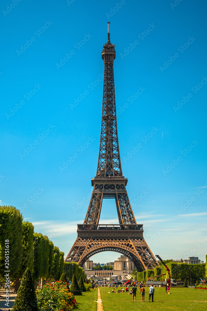 View of stunning Eiffel Tower, people and greenery under sunny blue sky, in Paris. Known as the “City of Light”, is one of the most impressive world’s cultural center. Northern France. Retouched photo
