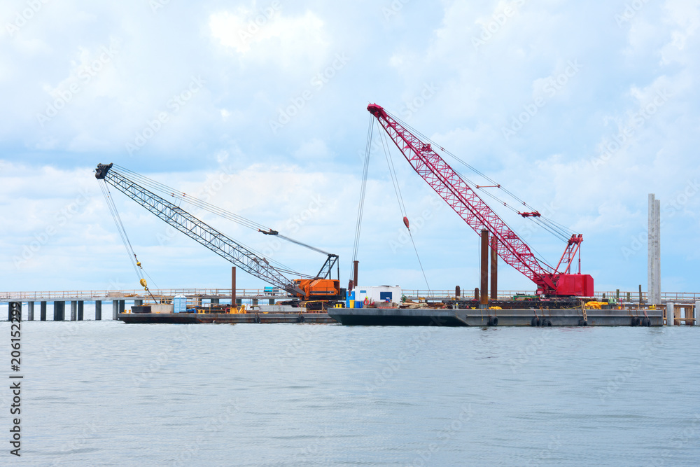 Bridge construction being done with two building crawler cranes on flat top boats floating on calm water in a bay or river.