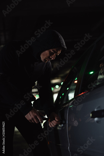 serious male robber in black hoodie intruding car by screwdriver
