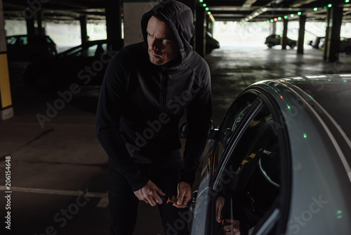 cautious male robber in black hoodie intruding car by screwdriver