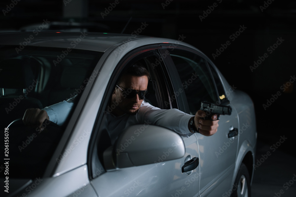 male private detective in sunglasses aiming by gun from car