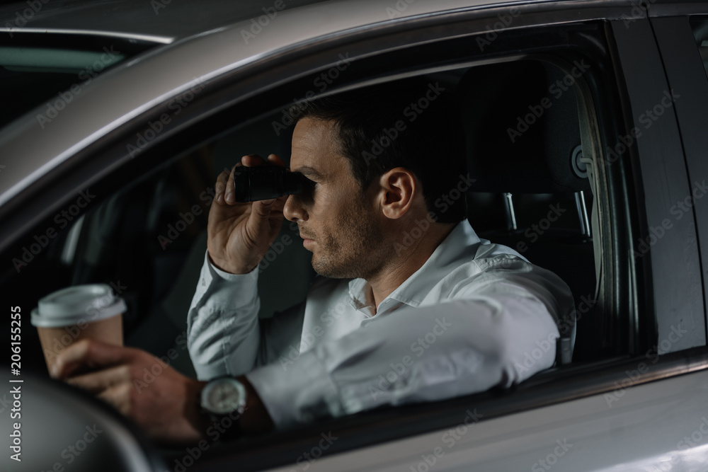 side view of undercover male agent doing surveillance by binoculars and drinking coffee in car