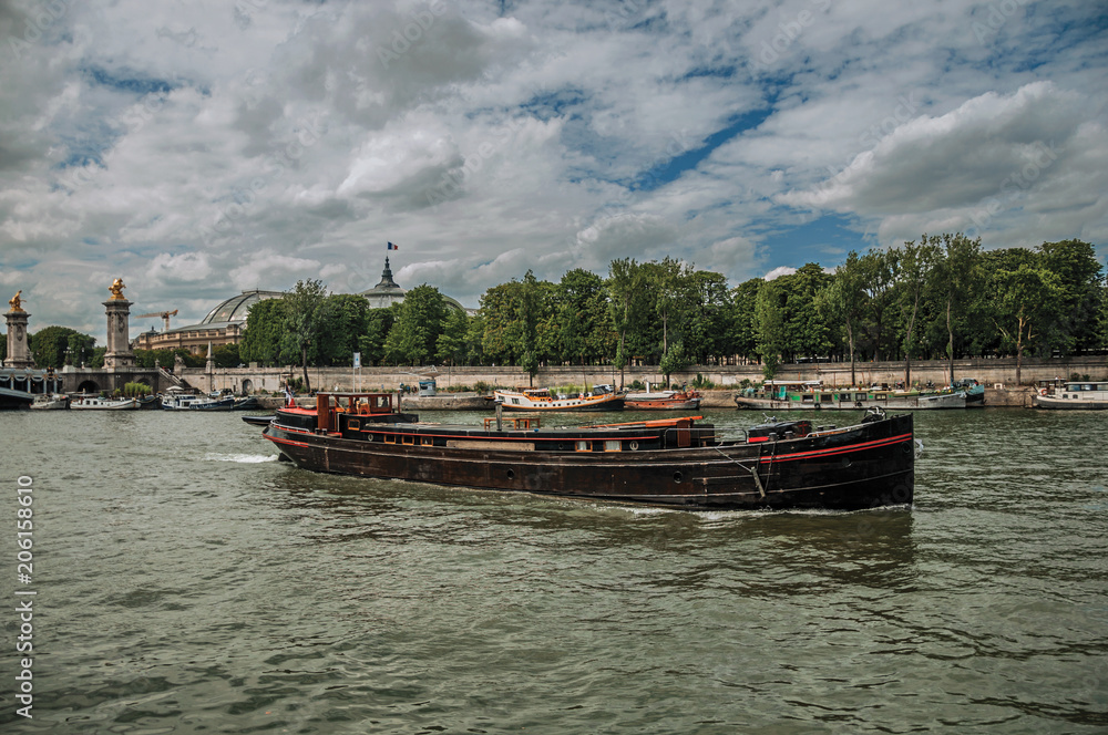 Big vessel and boats anchored at the Seine River bank with trees under a sunny blue sky in Paris. Known as the “City of Light”, is one of the most impressive world’s cultural center. Northern France.