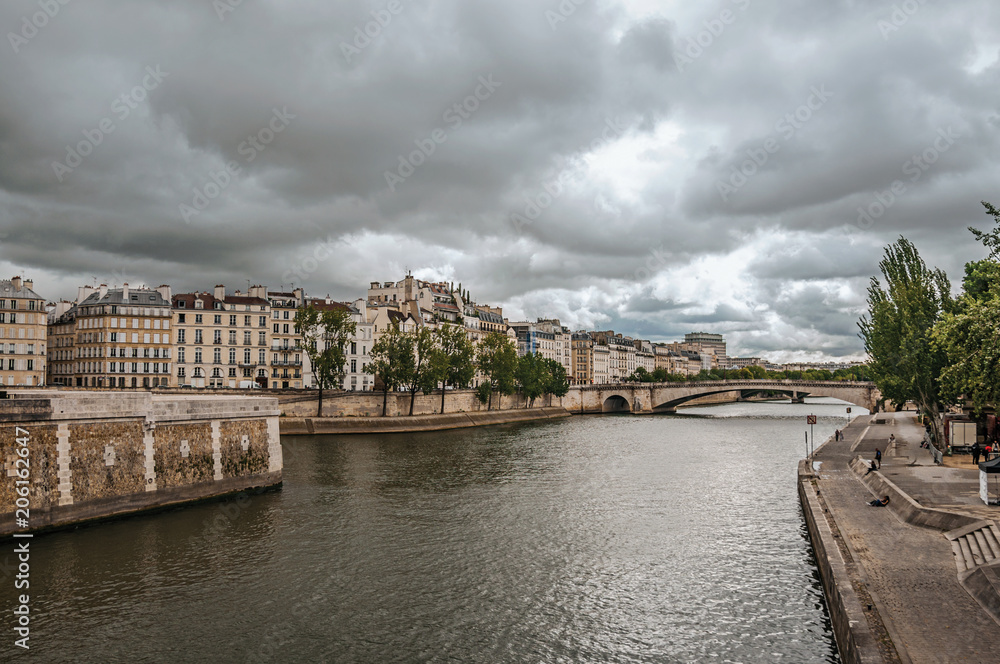 Old buildings, wall on the banks of the Seine River and bridge with cloudy sky in Paris. Known as the “City of Light”, is one of the most impressive world’s cultural center. Northern France.