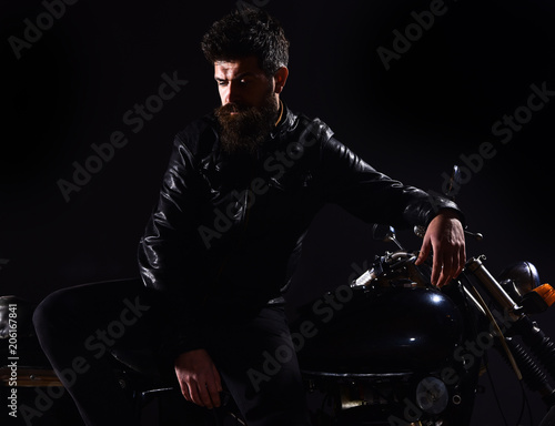 Macho, brutal biker in leather jacket stand near motorcycle at night time. Brutality and masculine concept. Man with beard, biker in leather jacket lean on motor bike in darkness, black background.