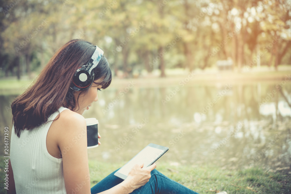 Obraz premium Beautiful girl is relaxing by listening to music. In the park there is a beautiful lake.She wears a white shirt And in her hands are choosing the song on the tablet. And she looks very happy.