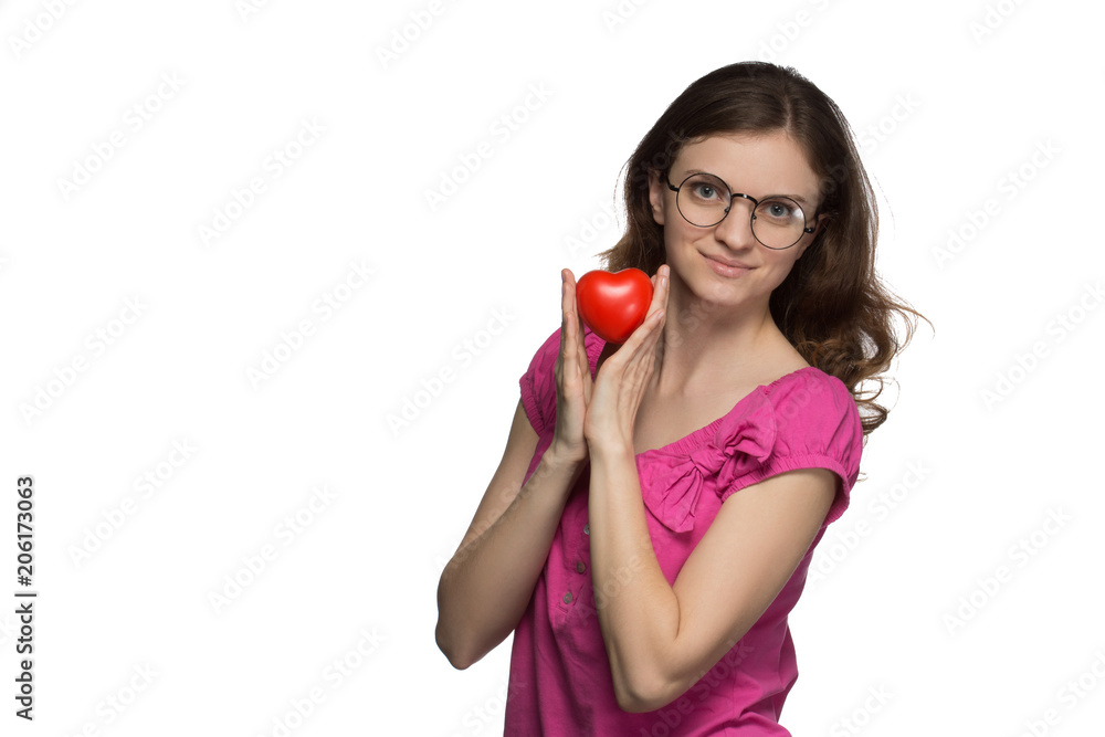 Beautiful brunette girl in glasses smiles and holds a heart in hands on a gray background Beautiful brunette girl in glasses smiles and holds a heart in hands on a gray background