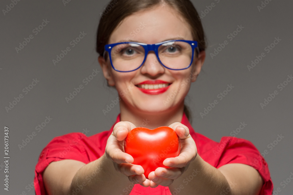 Beautiful brunette girl in glasses smiles and holds a heart in hands on a gray background Beautiful brunette girl in glasses smiles and holds a heart in hands on a gray background
