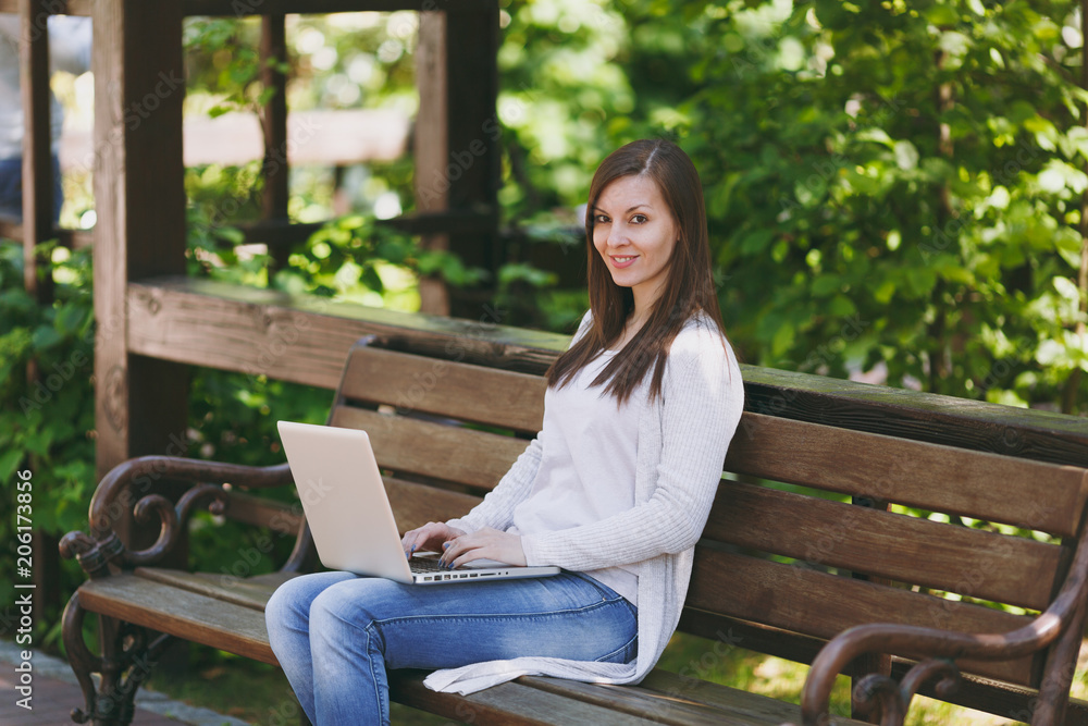 Young successful businesswoman in light casual clothes. Woman sitting on bench working on modern laptop pc computer in city park in street outdoors on nature. Mobile Office. Freelance business concept