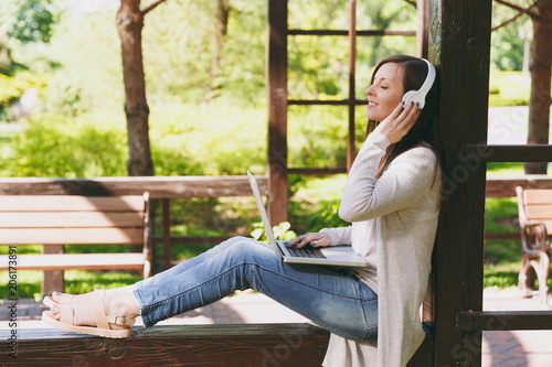 Free Download Young businesswoman in light casual clothes. Woman working on modern laptop pc computer, listen music in headphones on head in street outdoors. Mobile Office. Freelance business concept. Side view. - GFXLoad
