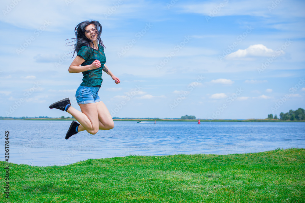 Beautiful funny smiling young woman jumping, against background of blue sky and green park. Concept of active lifestyle 