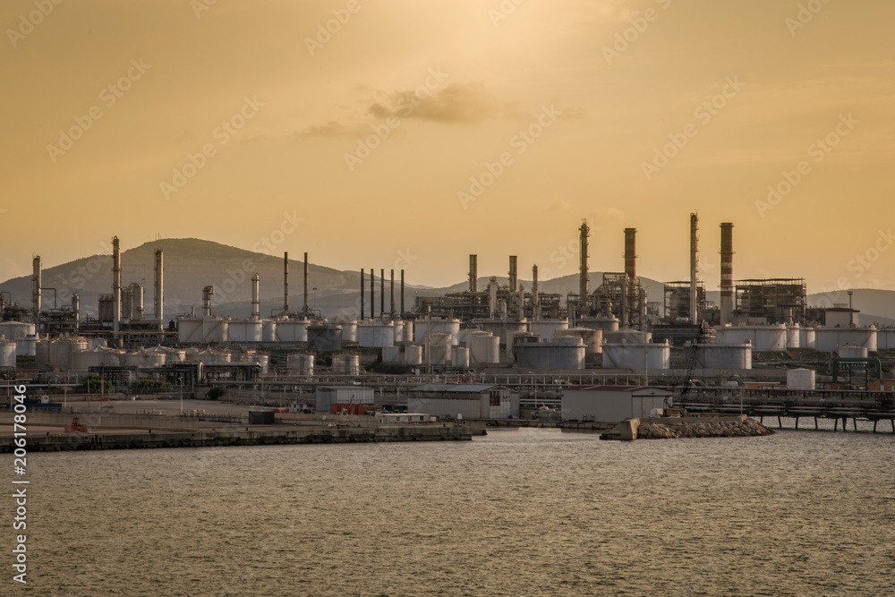 Skyline industrial area Cagliari, Sardinia Italy. View from the sea of an oil refinery plant from industry zone with cloudy sky at sunset. Oil storage tank in port area.