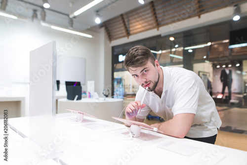 Handsome man stands next to a showcase with tablet stylus in his hands and looks at the camera. Buyer looks at the tablets in the electronics store.