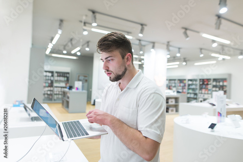 Handsome man uses a notebook in a light tech store. A buyer chooses a notebook in a modern electronics store. Buy a laptop.