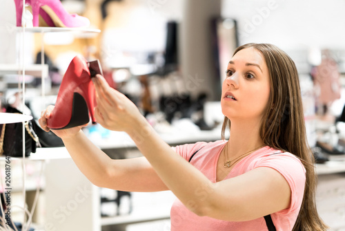 Shocked woman looking at price tag of too expensive shoes in fashion store while shopping. Unhappy customer holding high heels. Sad shopper with overpriced product.