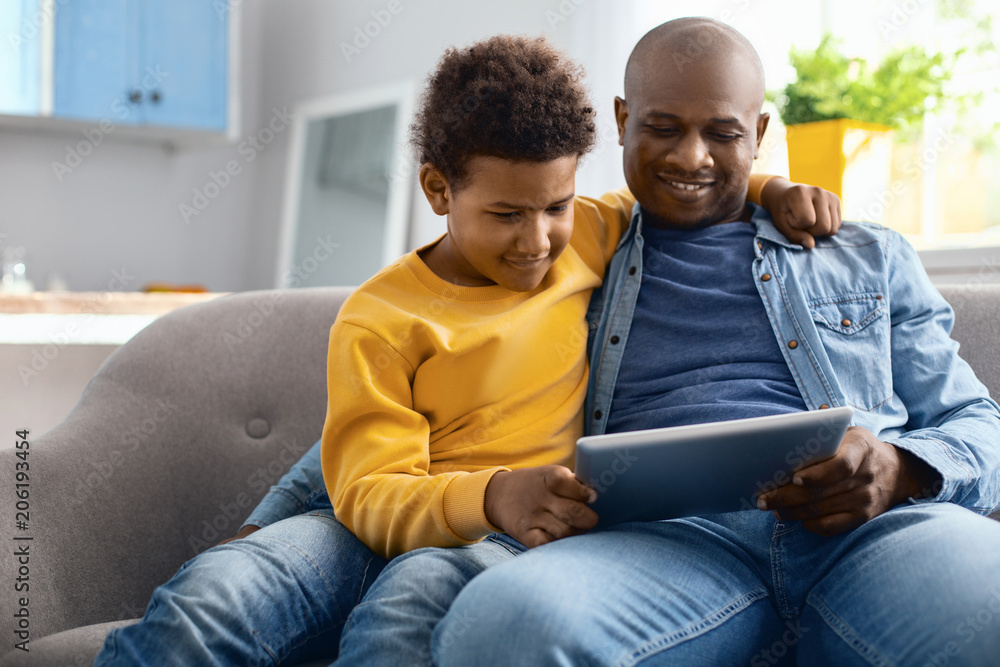 Funny videos. Pleasant young father and his pre-teen son sitting on the sofa and watching videos on tablet together while the boy hugging his father Funny videos. Pleasant young father and his pre-teen son sitting on the sofa and watching videos on tablet together while the boy hugging his father