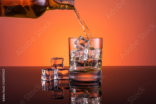 Whisky alcohol pouring into glass with ice drink on warm dark orange background