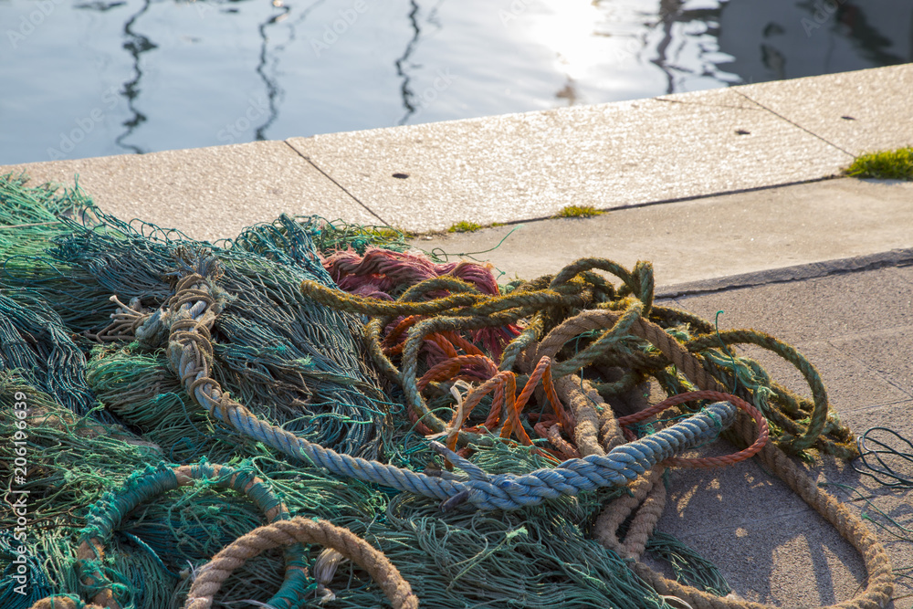 Pile of fishing nets with their associated ropes lying on a quay in a fishing harbour with sunlit water in the background