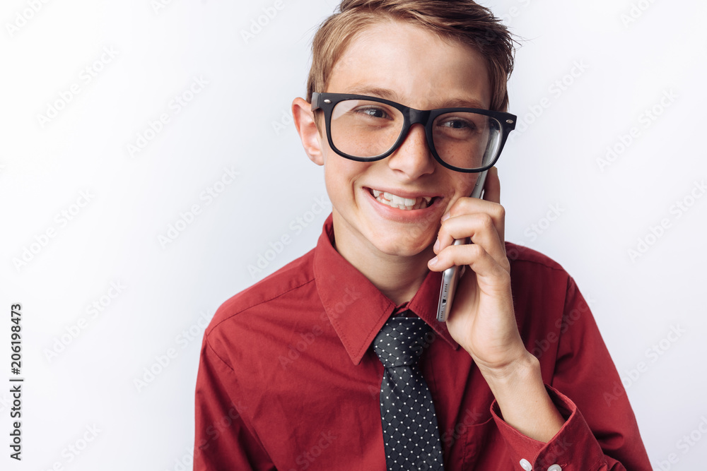 Portrait of positive and emotional schoolboy talking on phone, white background, glasses, red shirt, business theme, advertising,