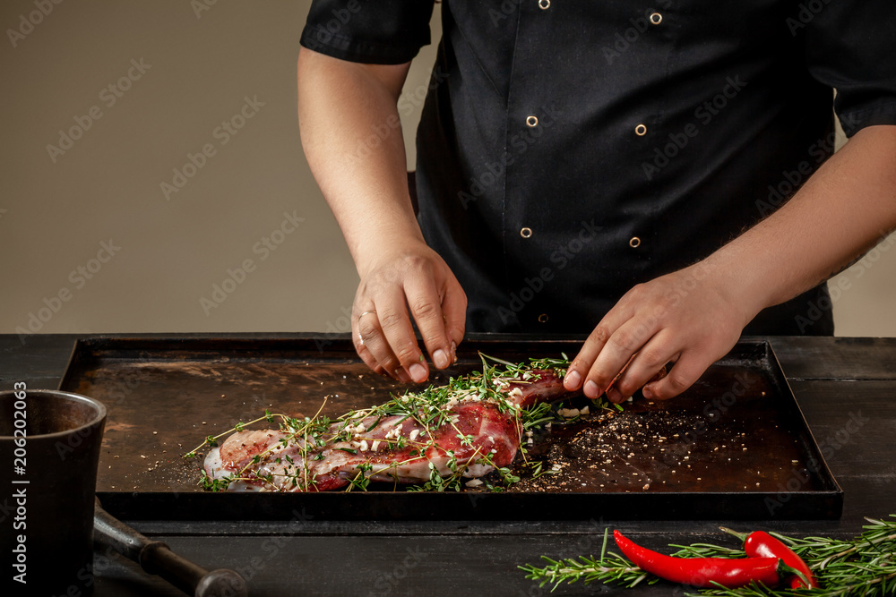 Male chef rubbing raw lamb shanks with greens and spices on stone tray on wooden table. Chef cooking appetizing shank of lamb.