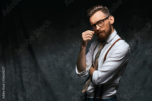 Young stylish hipster with cool hairstyle and beard dressed in white shirt and suspenders is thinking of a new creative idea looking at viewer