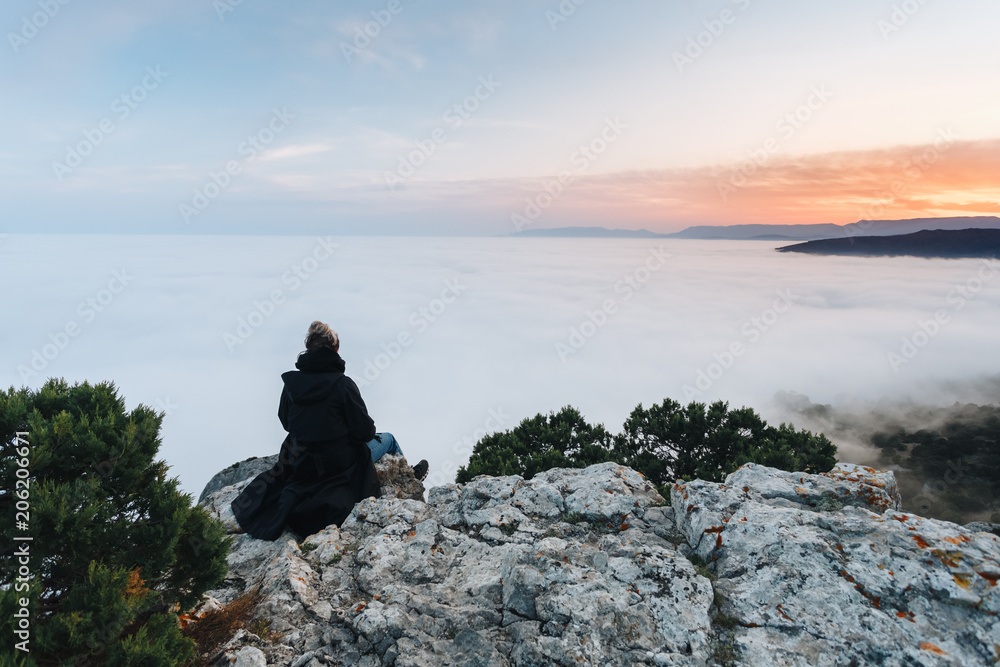 The girl sits on the edge of the cliff above the dense fog that covered the sea, on Mount Kosmos in the Crimea