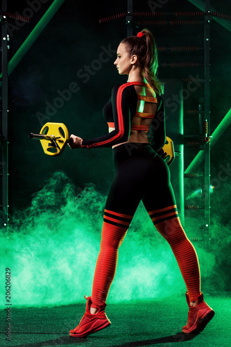 Fototapeta Naklejka Na Ścianę i Meble -  Young woman in the gym with a barbell, in a beautiful sportswear, against a dark background with color lights.