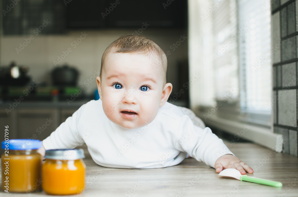 charming child with blue eyes on the table crawling to the jars of fruit puree charming child with blue eyes on the table crawling to the jars of fruit puree