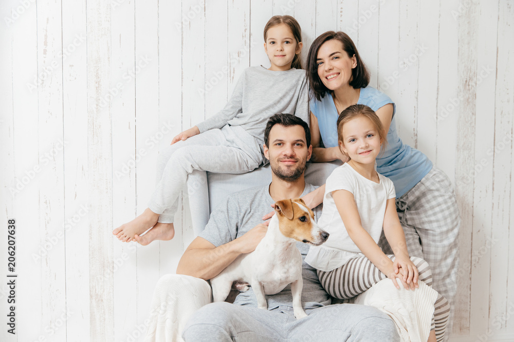 Family portrait. Happy parents with their two daughters and dog pose together against white background, spend free time at home, being in good mood. Mother, father and small sisters pose indoor