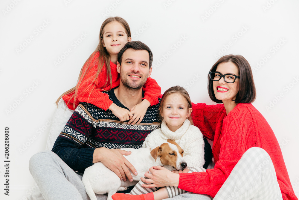Portrait of happy family members with positive expressions, cuddle and support each other, have good relationship. Delighted father, mother, two sisters and small puppy pose together indoor.