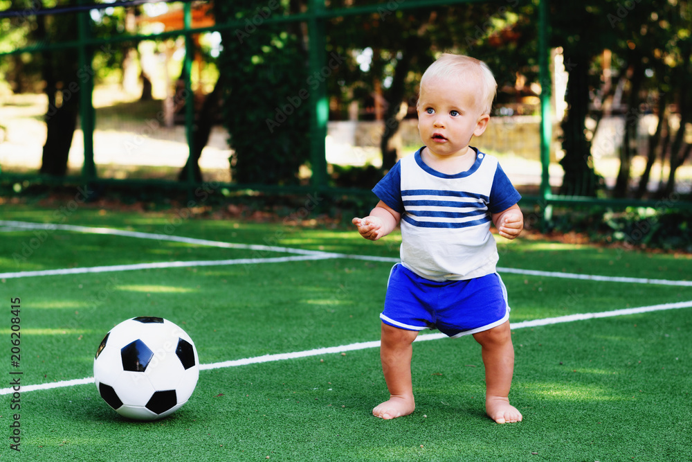 Little boy in blue shorts playing with soccer ball at sports ground. Blonde child in sports uniform standing at football field with a ball. Summer kids activity concept